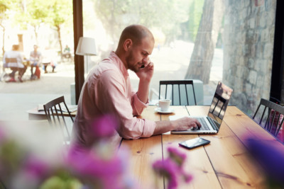 A man works in a coffee shop while talking on the phone